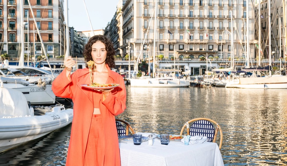 Model im orangefarbenen Kleid am Hafen mit Yachten, Drykorn-Kampagne FS/SS 23 in Neapel