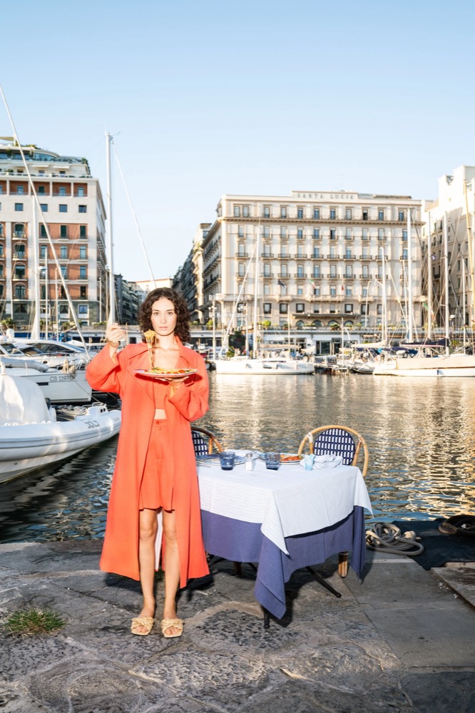 Model im orangefarbenen Kleid neben einem Tisch am Hafen, Drykorn-Kampagne FS/SS 23 in Neapel