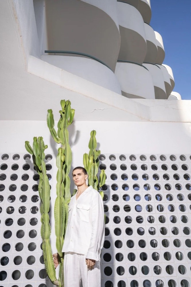 Model in Weiß neben einem Kaktus vor einer durchbrochenen Mauer, Drykorn-Kampagne FS/SS 24 Teil II auf Mallorca