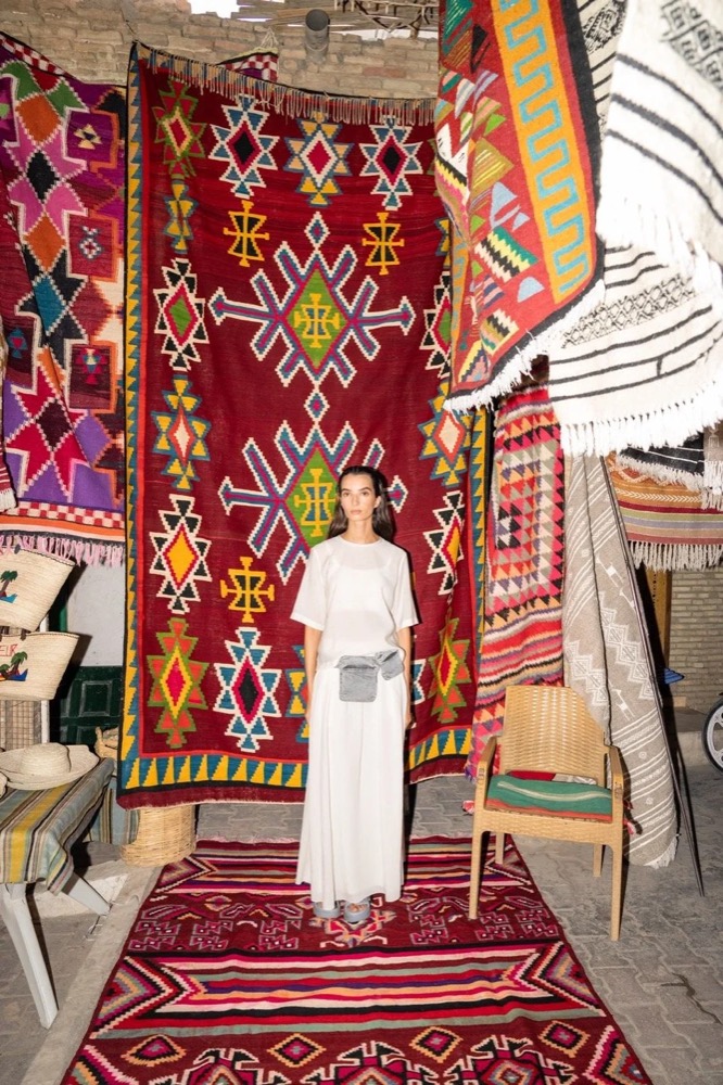 Model in Weiß vor hängenden Teppichen in einem Souk, Drykorn-Kampagne FS/SS 24 in Tozeur, Tunesien