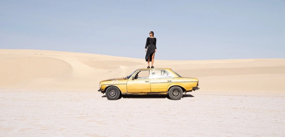 Model steht auf einem gelben Auto in der Wüste, Drykorn-Kampagne FS/SS 24 in Tozeur, Tunesien