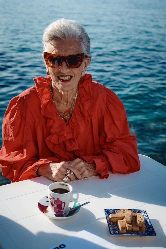 Ältere Frau in orangefarbener Bluse und Sonnenbrille am Frühstückstisch am Meer, Drykorn-Kampagne FS/SS26 in Genua