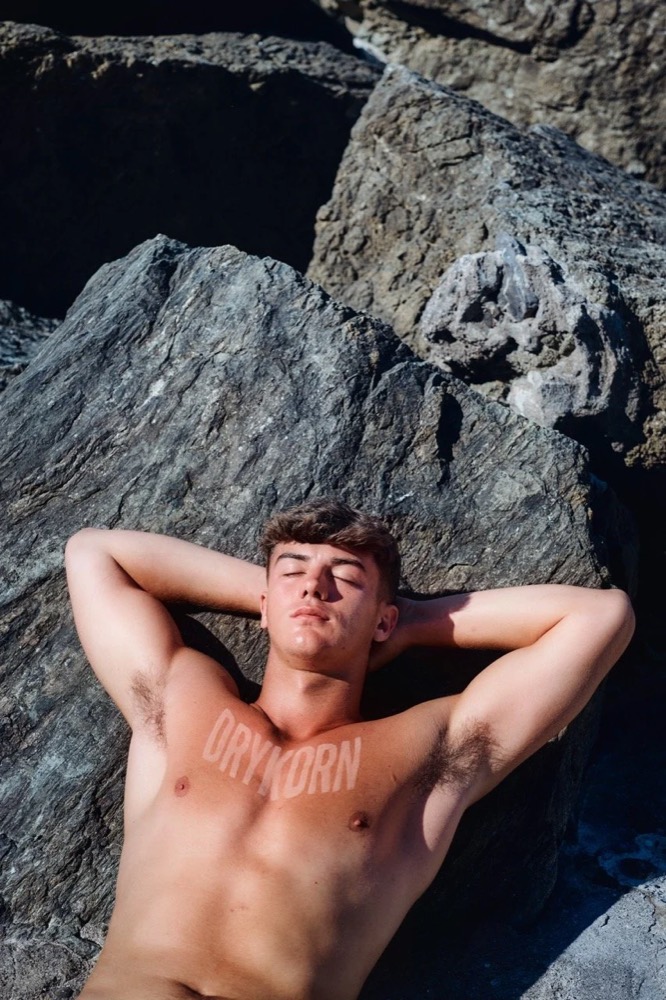 Model mit nacktem Oberkörper auf dunklen Felsen liegend, Drykorn-Kampagne FS/SS26 in Genua