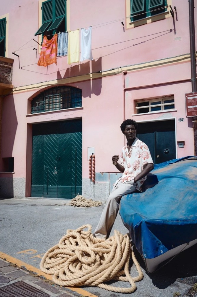 Model sitzt auf einem blauen Boot vor einem rosa Haus, Drykorn-Kampagne FS/SS26 in Genua