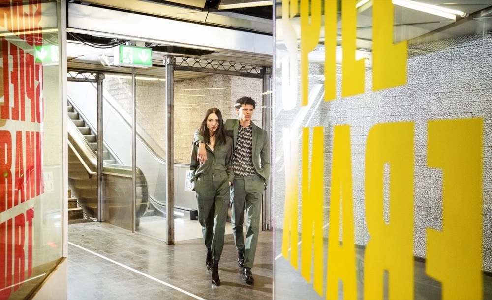 Zwei Models in einer Bahnhofspassage mit gelben Plakaten, Drykorn-Kampagne HW/AW 2021 in Frankfurt