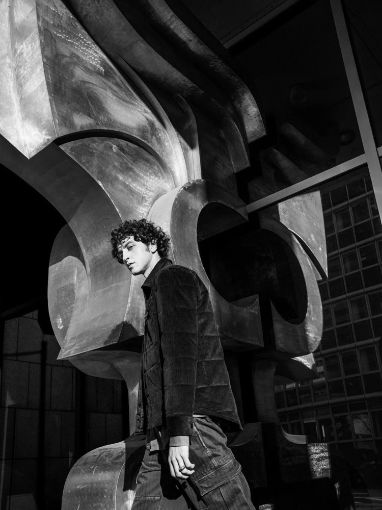 Schwarzweißaufnahme eines Models unter einer modernen Skulptur, Drykorn-Kampagne HW/AW 2021 in Frankfurt