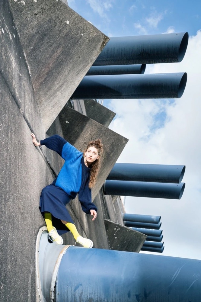 Model in blauem Strick und weiter Hose auf brutalistischem Beton mit Rohren, Untersicht, Drykorn-Kampagne HW/AW 22