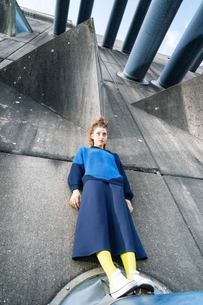Model im blauen Pullover und gelben Stiefeln unter Betonrohren, Drykorn-Kampagne HW/AW 22