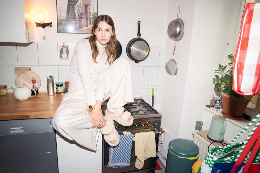 Model in Weiß sitzt in einer vollgestellten Küche, Drykorn-Kampagne HW/AW 22
