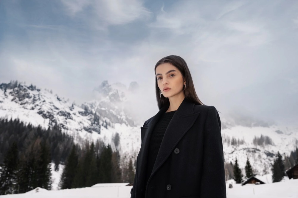 Model im schwarzen Mantel in einer verschneiten Berglandschaft, Drykorn-Kampagne HW/AW 23 in Bad Gastein