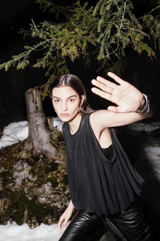 Model in Schwarz hebt die Hand unter einer Tanne, Drykorn-Kampagne HW/AW 23 in Bad Gastein