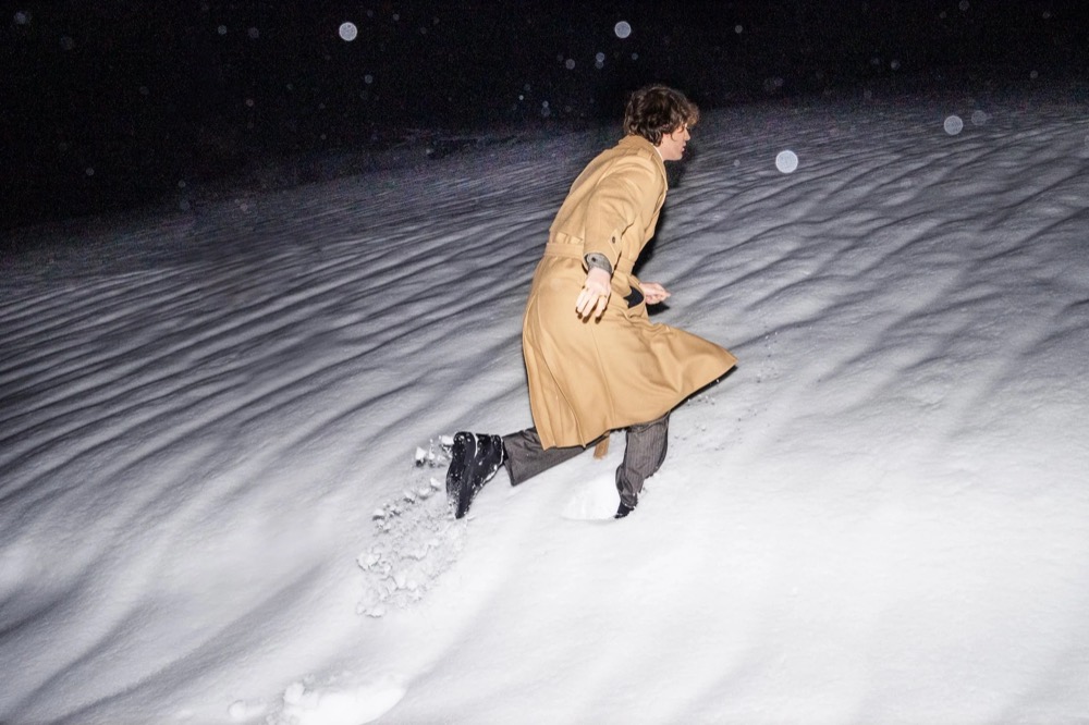 Model im kamelfarbenen Mantel nachts im Schnee, Drykorn-Kampagne HW/AW 23 in Bad Gastein