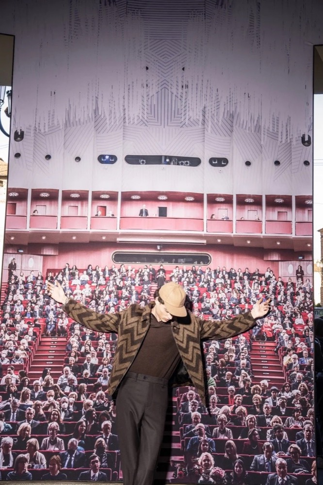 Model mit ausgebreiteten Armen vor einer Wand aus Sitzplatzfotos, Drykorn-Kampagne HW/AW 24 in einem Theater in Turin