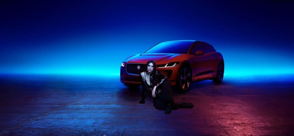 Dua Lipa sitzt unter blauem Neonlicht vor einem roten Jaguar-SUV, Jaguar The Pace