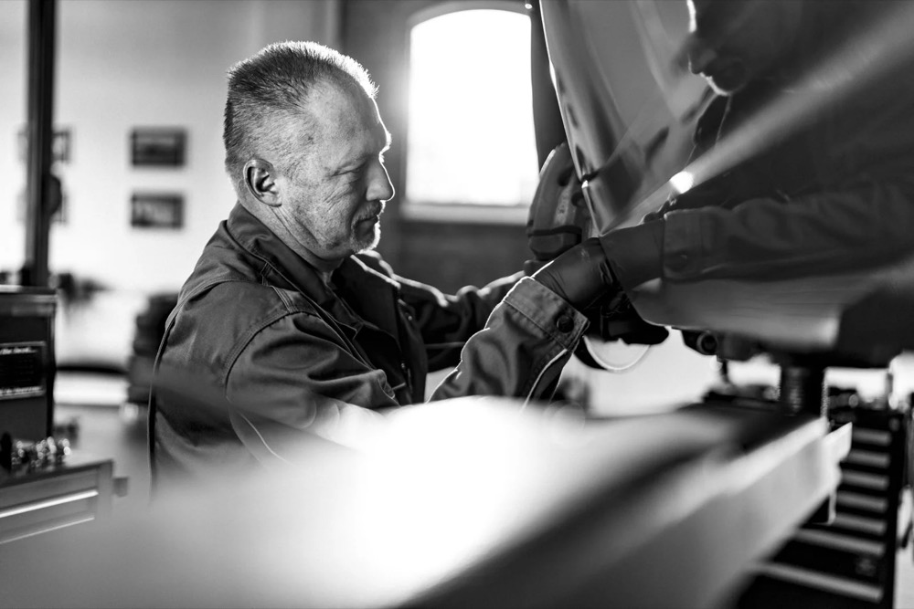 Schwarzweißaufnahme eines Mechanikers, der unter einer Motorhaube arbeitet, Jaguar Land Rover Tradeparts