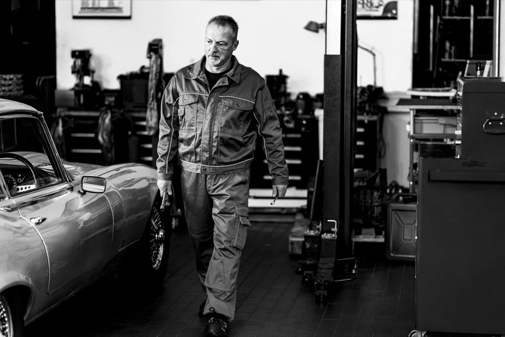 Mechaniker geht in der Werkstatt an einem klassischen Jaguar vorbei, Jaguar Land Rover Tradeparts