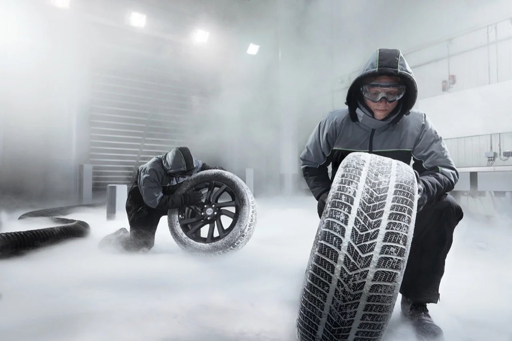 Mechaniker in Kapuzenjacke kniet mit einem Winterreifen im Nebel, Jaguar Land Rover Winterräder und -reifen