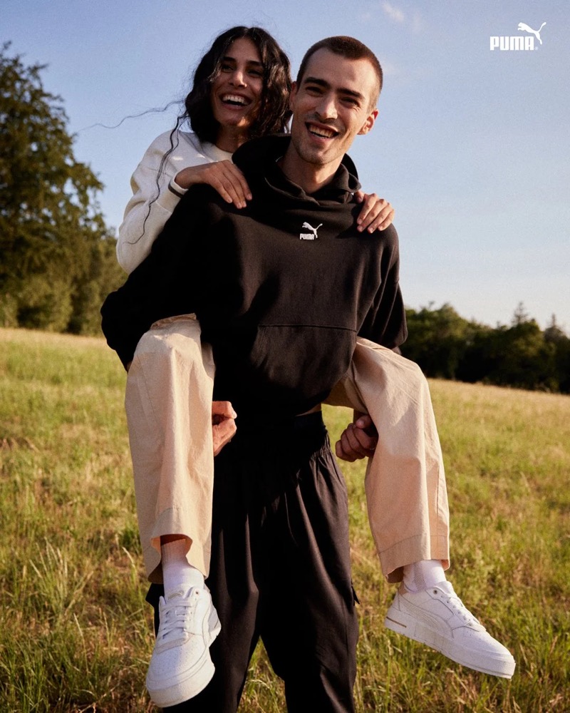 Model nimmt ein anderes Model auf einer Wiese huckepack, Puma AW23