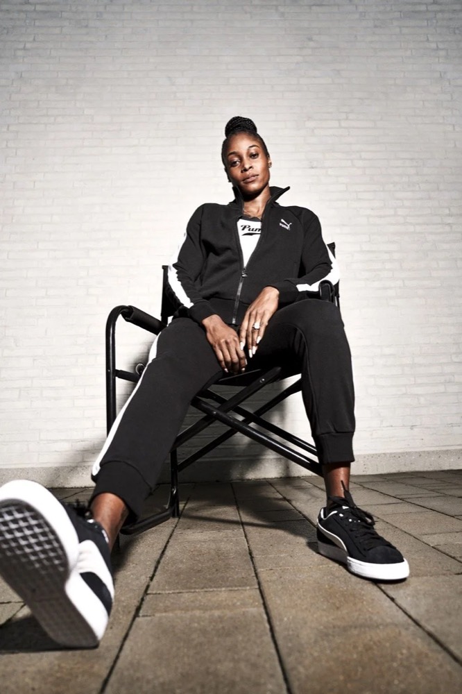 Elaine Thompson-Herah im schwarzen Puma-Trainingsanzug auf einem Stuhl