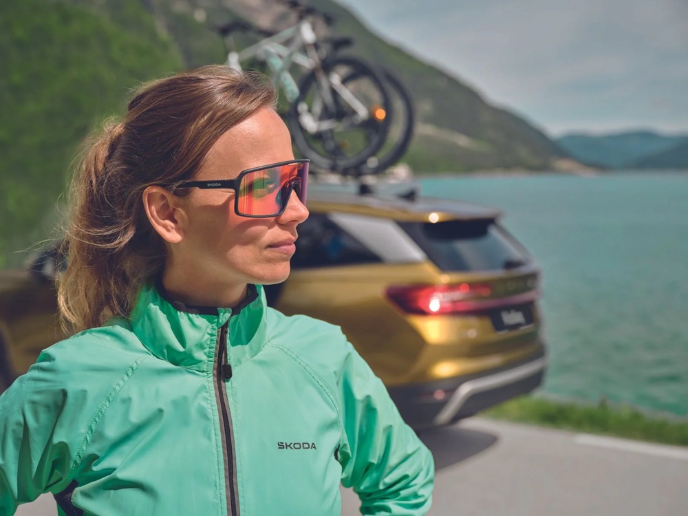 Radfahrer in grüner Jacke neben einem Škoda Kodiaq mit Fahrradträger an einem See, Škoda-Zubehörkampagne
