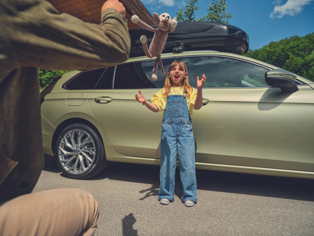 Mädchen in Latzhose neben einem Škoda Superb mit offenem Kofferraum, Škoda-Zubehörkampagne